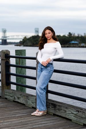 Woman in white off-shoulder top and jeans standing on wooden pier with bridge and water in background.