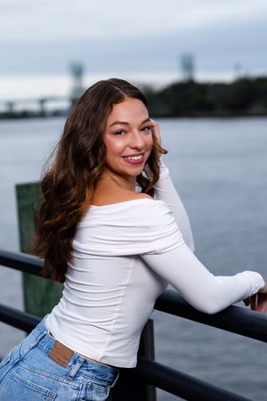 Someone wearing a white long sleeve top and blue jeans poses playfully by a waterfront railing at dusk.