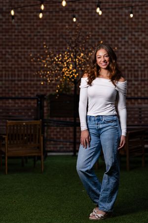 Stylish casual outfit with off-shoulder white top and jeans against brick wall with twinkle lights in background.