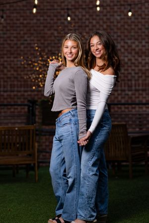 Two friends strike a playful pose together against a brick wall with string lights in the background.