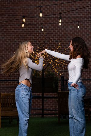 Two friends playfully dancing together at dusk against a brick wall with twinkling lights in the background.