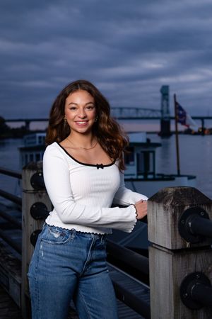 Portrait of a student in a white sweater and jeans standing by a riverside with a bridge in the background at dusk.