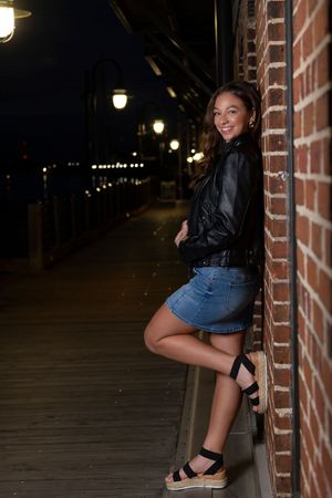 Person in leather jacket and denim skirt poses against brick wall on boardwalk at night with illuminated lamps.