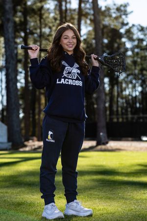 Person wearing navy blue lacrosse sweatshirt and white sneakers poses on grassy field with tall trees in background.