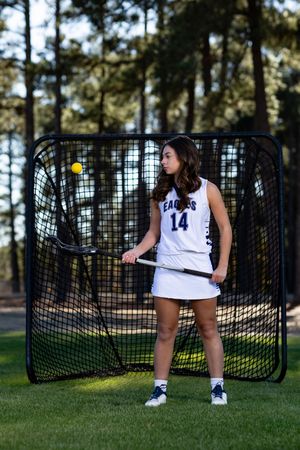 A high school lacrosse player stands in front of a training net in uniform number 14 on an outdoor field.