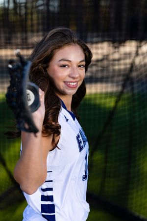 Softball player in a white uniform smiles and poses for a portrait series on a field.
