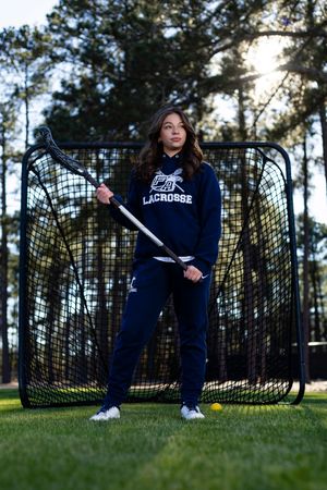 Lacrosse player stands with stick in front of practice net on grass field at sunset with pine trees in background.