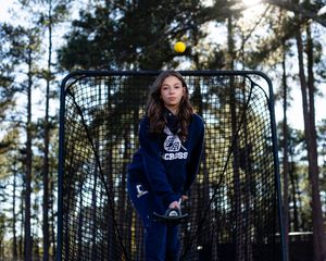A player stands in front of a lacrosse goal net surrounded by tall pine trees at dusk.