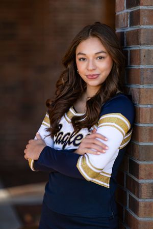 Student in navy and gold spirit wear cheerleading uniform poses against brick wall for portrait.