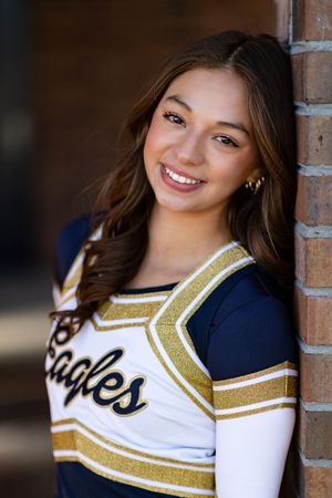 A cheerleader in a navy and white uniform with gold trim poses for a portrait against a brick wall backdrop.