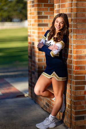 A cheerleader in a navy and gold uniform poses against a brick pillar on a sunny day.
