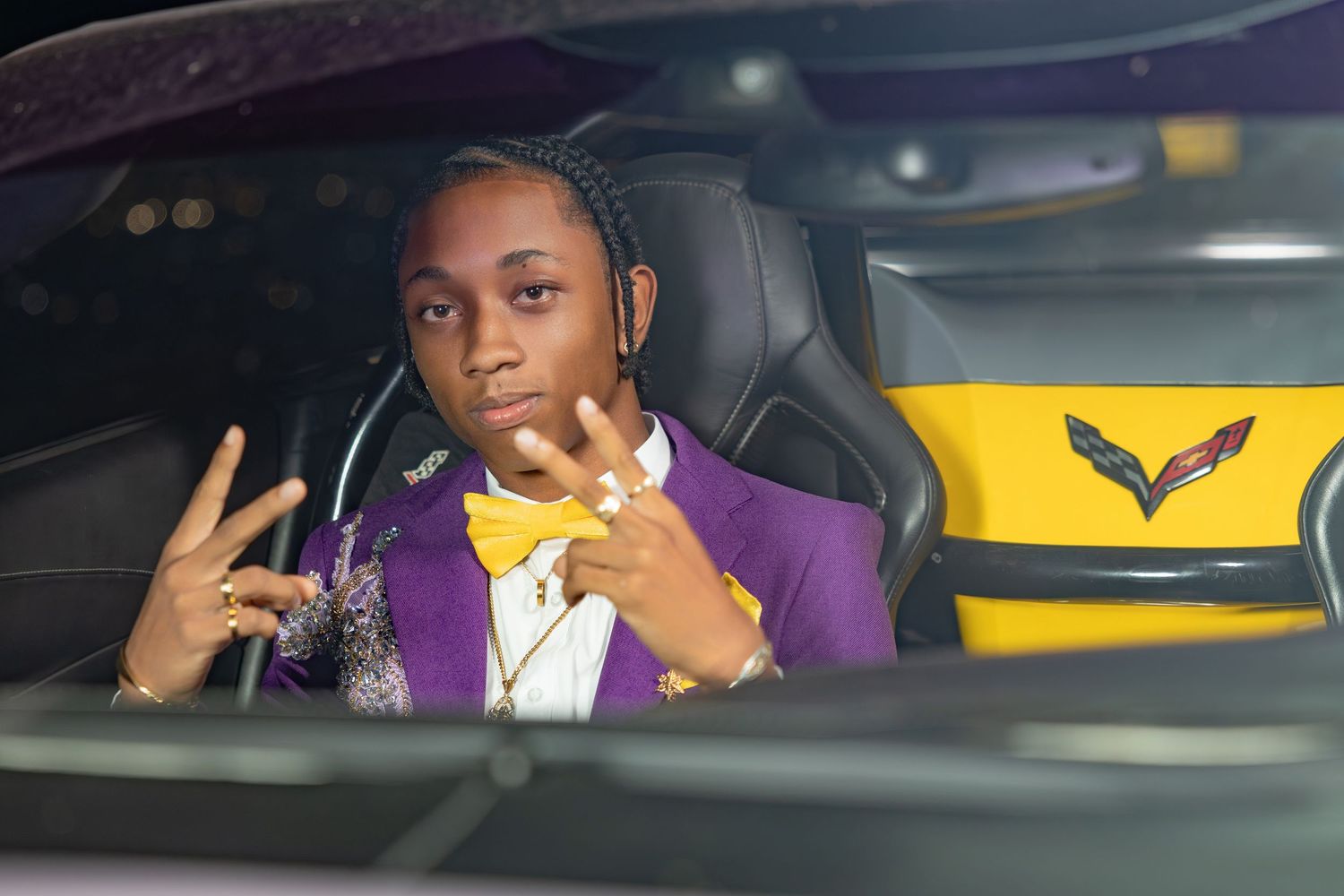 Chesson Liburd poses during his prom night portrait session, dressed in a purple suit with a yellow bowtie, sitting in a stylish car.