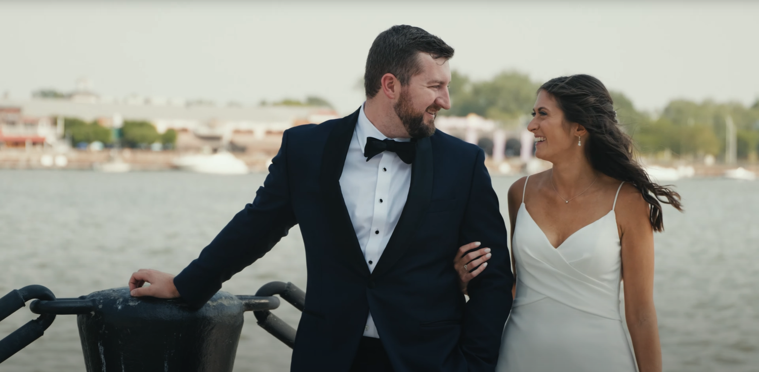 A couple in wedding attire share a moment together while standing near a waterfront.