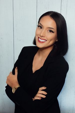 Professional portrait against white wooden wall shows confident smile and black blazer with arms crossed.