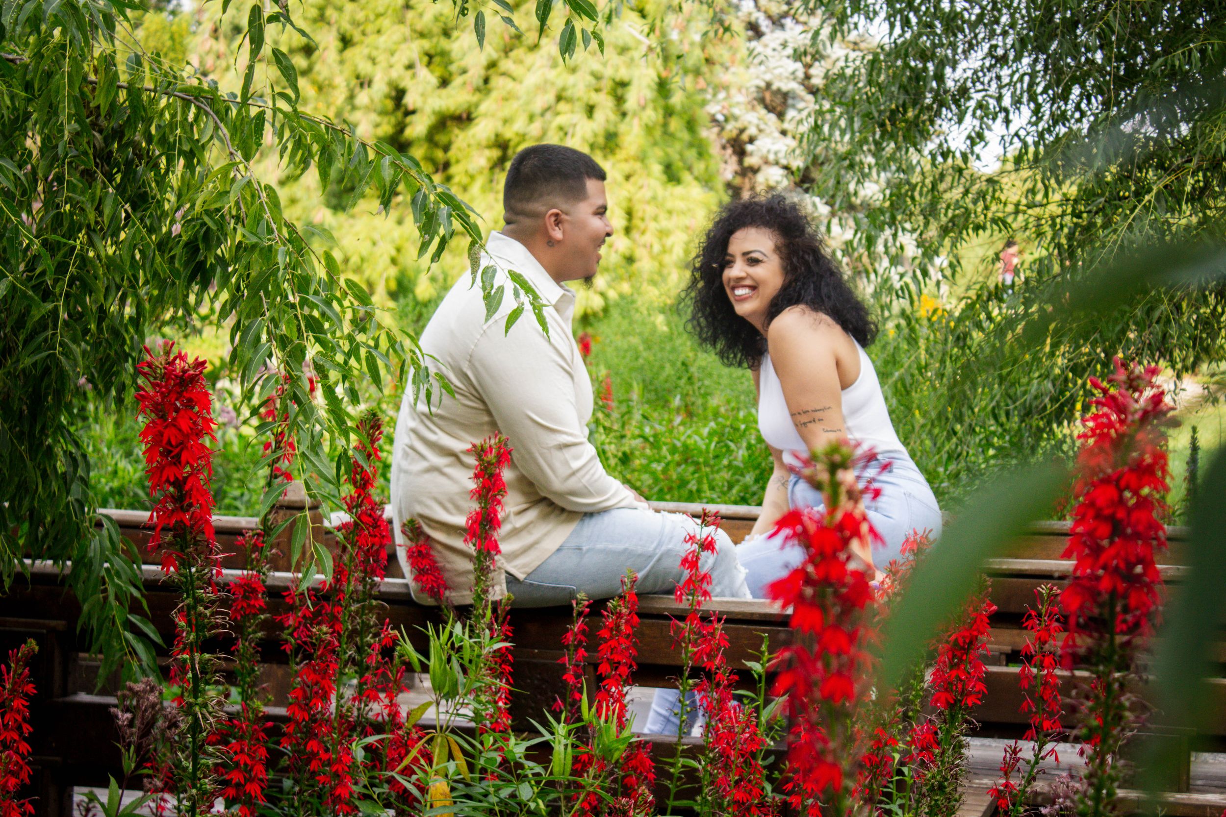 A romantic couple share a joyful moment in a garden surrounded by vibrant red flowers and lush greenery.