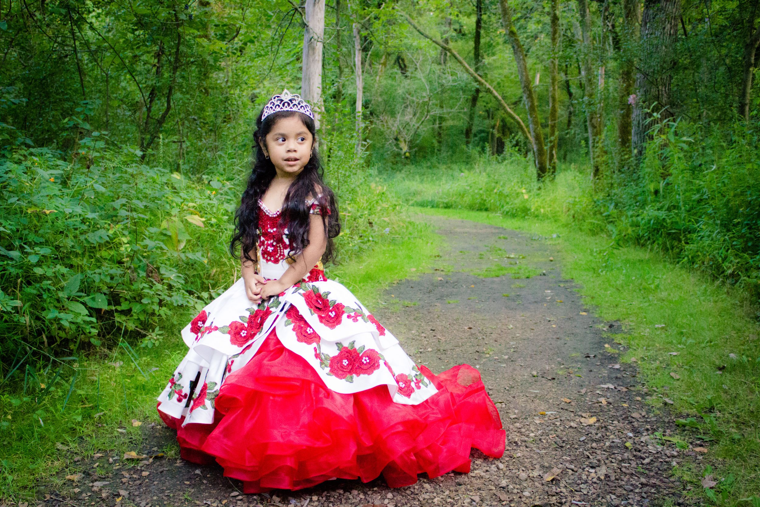 A person in an elegant red and white lace dress poses along a peaceful forest trail surrounded by lush greenery.