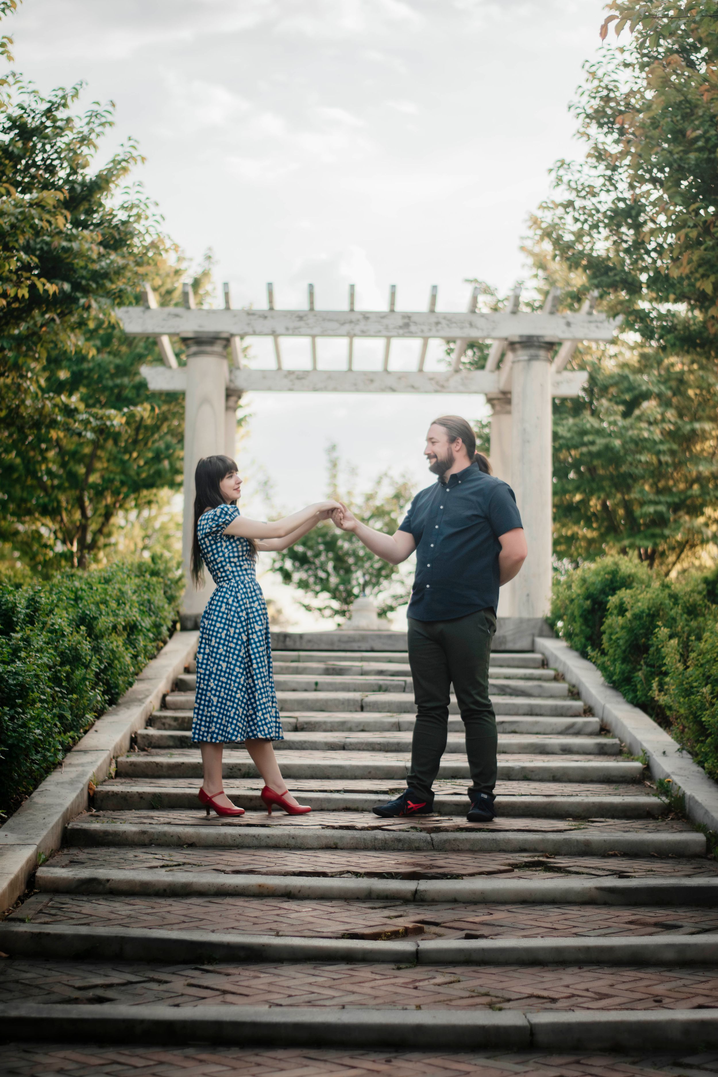Two figures stand together on rustic wooden steps under a white garden pergola surrounded by greenery.