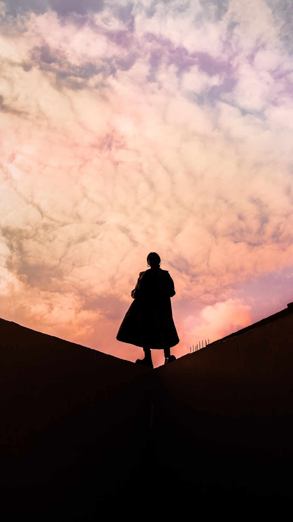 A silhouette of a person walking along a the edge of a high wall against a dramatic pink and orange sunset sky with clouds.