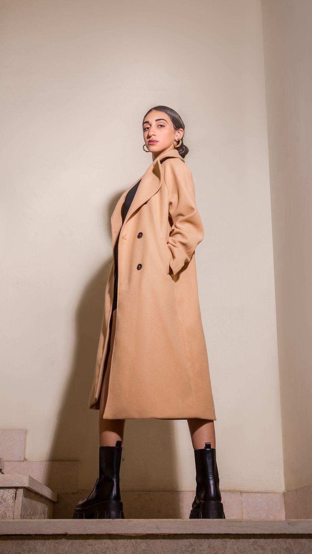 Model wearing a beige wool coat with black boots poses in a minimalist studio setting against white walls.