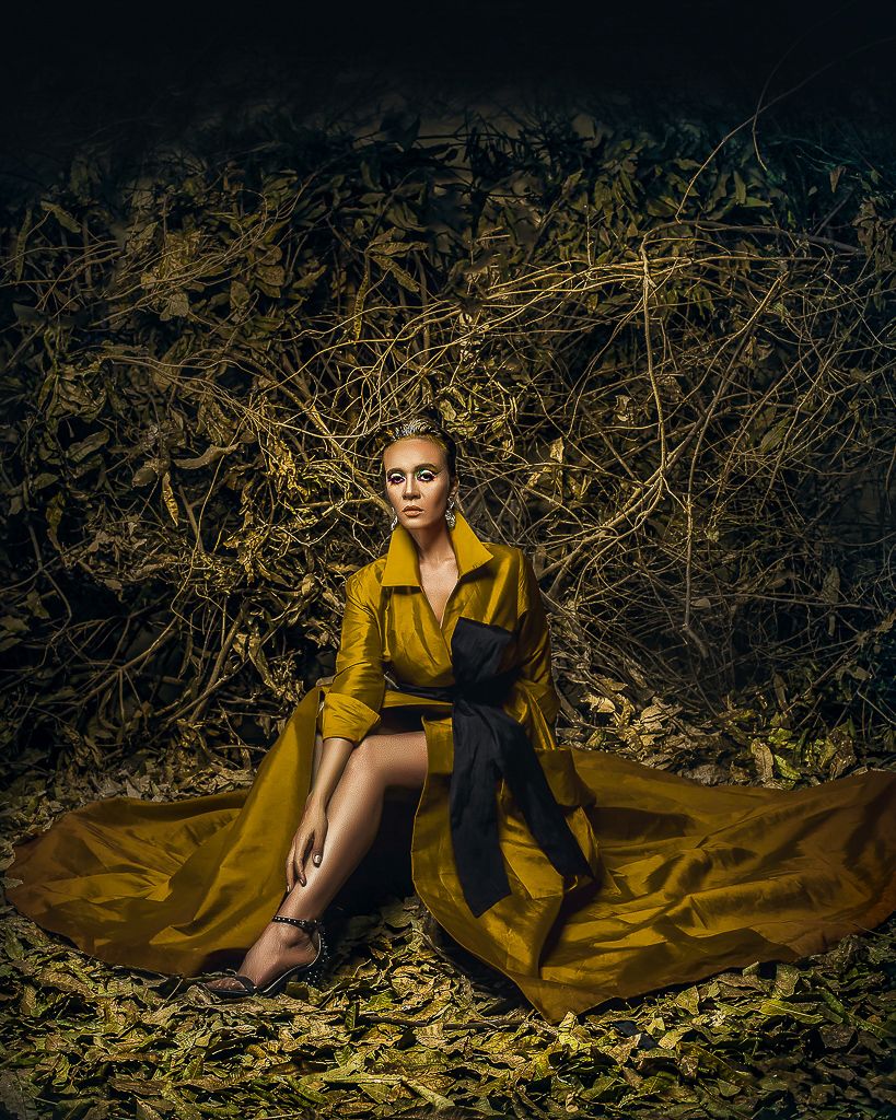 Fashion model in flowing yellow dress poses dramatically against dark foliage background in artistic photoshoot series.