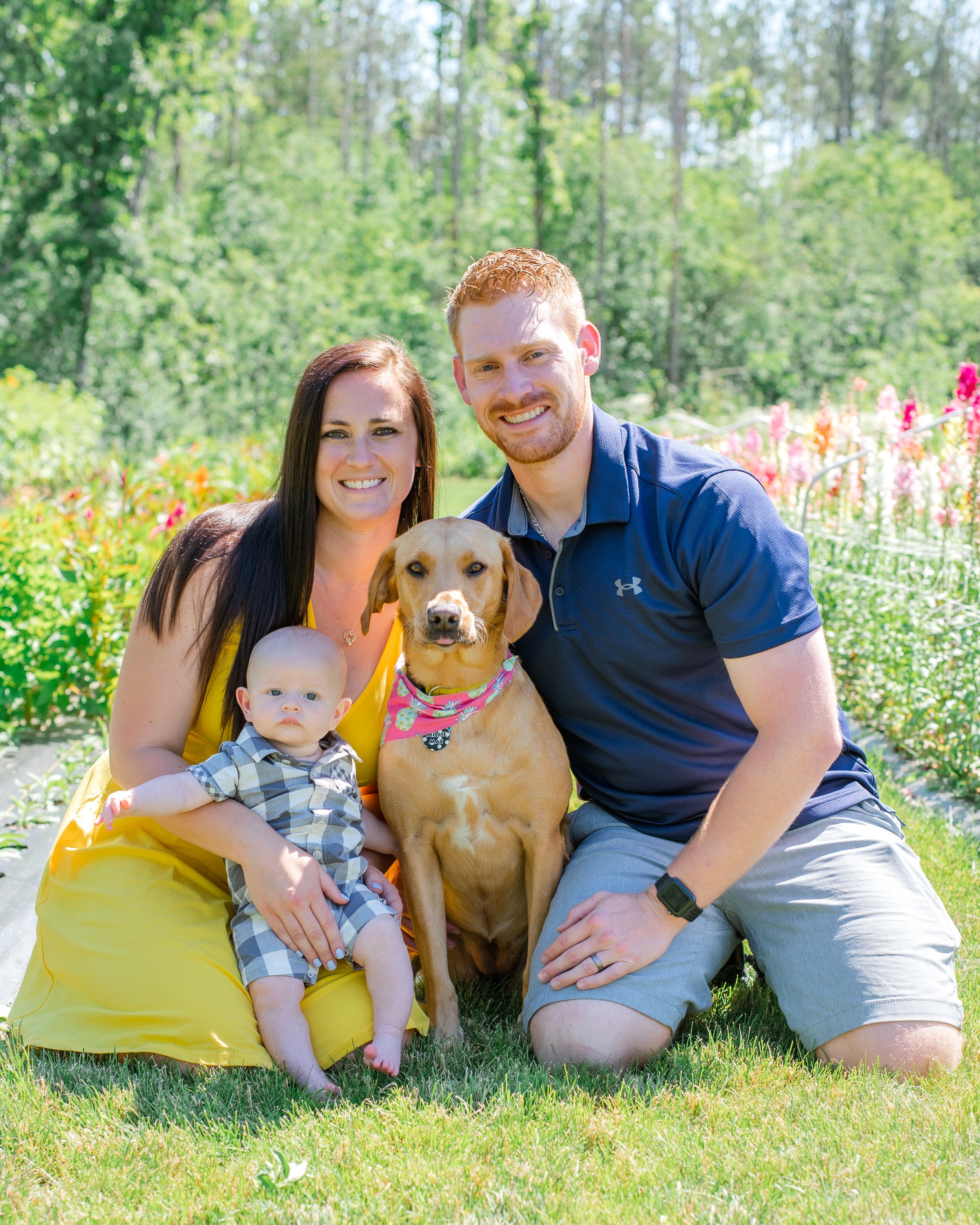 A family and their brown dog pose together on grass with colorful flowers blooming in the background.