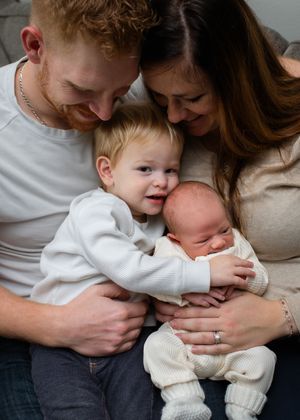 Loving family embraces on couch while young child holds newborn baby sibling in white sweater.