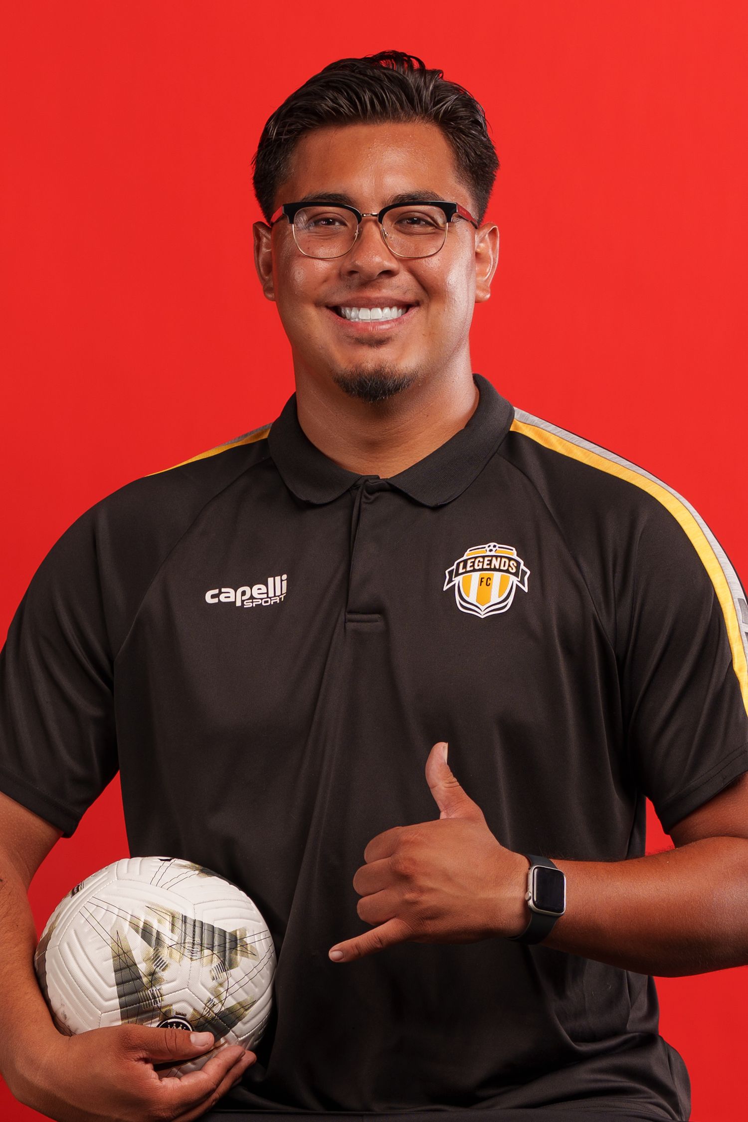 Soccer coach in black uniform holding volleyball and making shaka sign against red background.