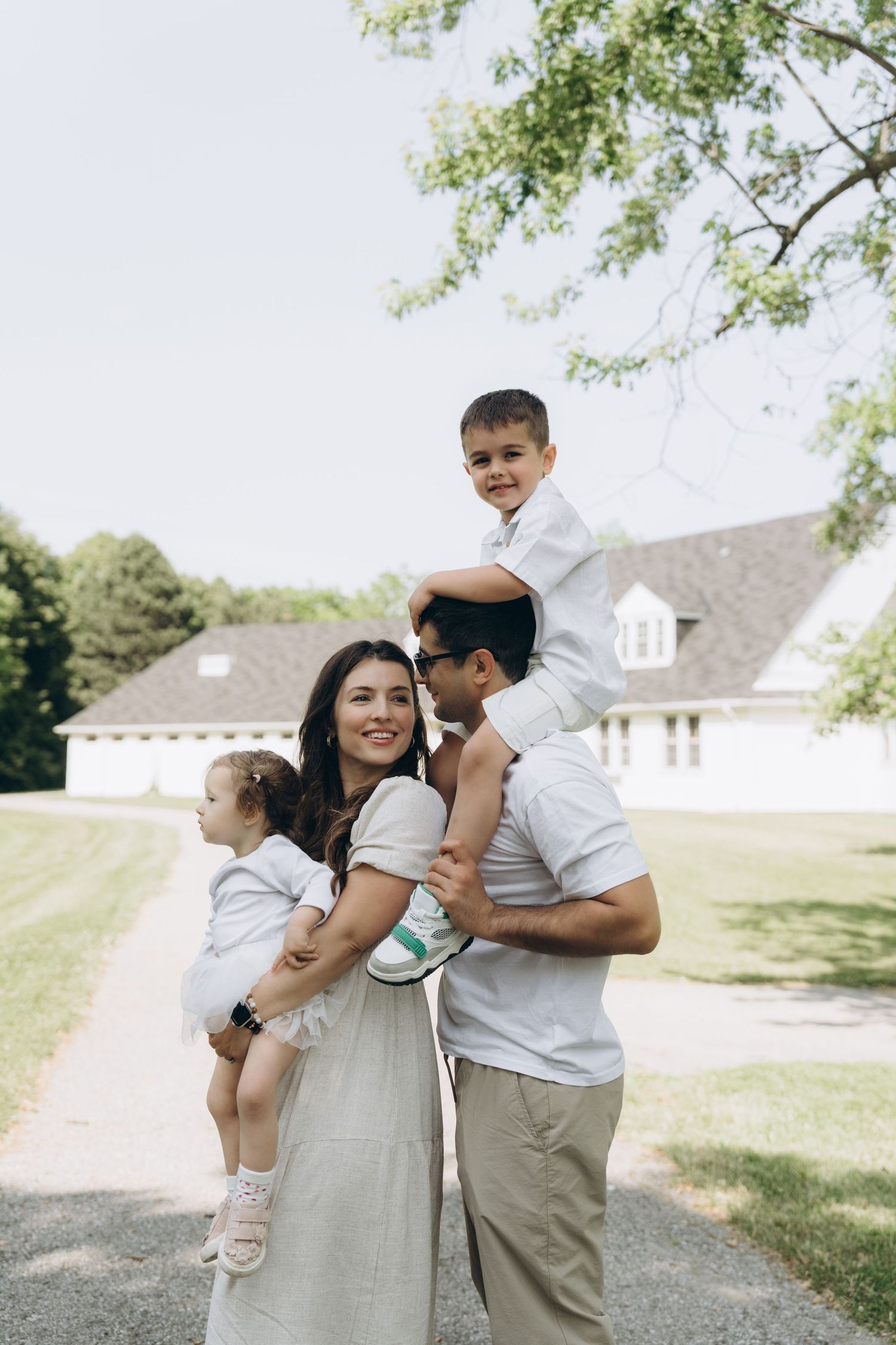 A family wearing white and grey clothing poses together outdoors on a sunny day near a house with trees.