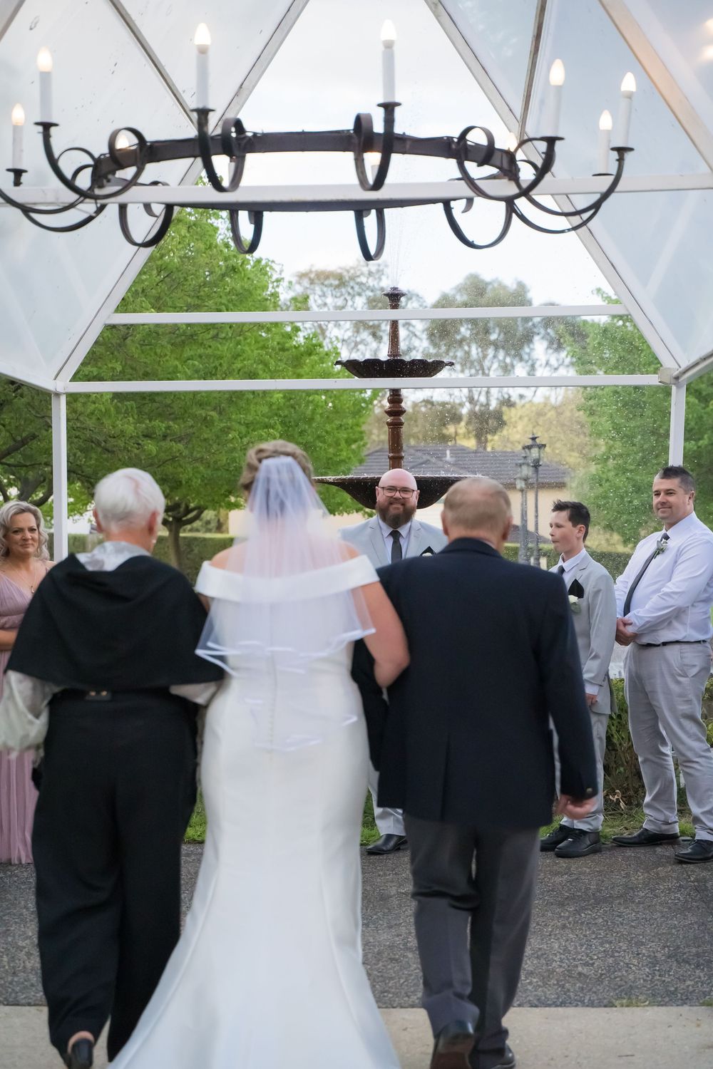 Wedding guests gather under a clear tent decorated with chandeliers during an outdoor celebration.