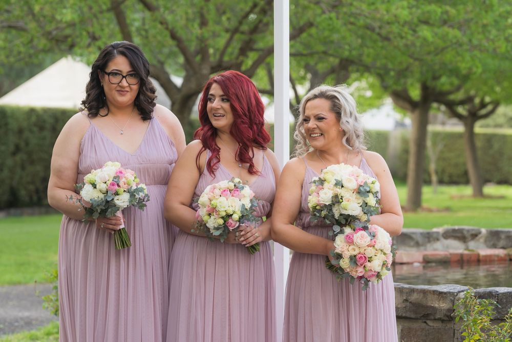 Bridesmaids in dusty rose dresses holding pink and white floral bouquets smile together at an outdoor wedding venue.