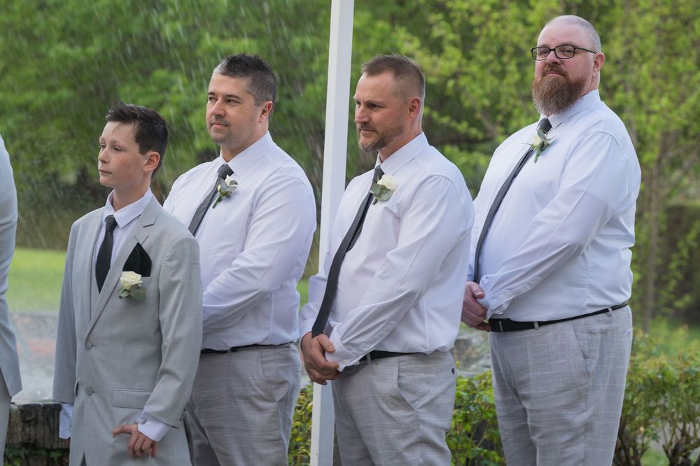 Wedding party groomsmen in gray suits and white shirts line up outdoors in a natural setting.