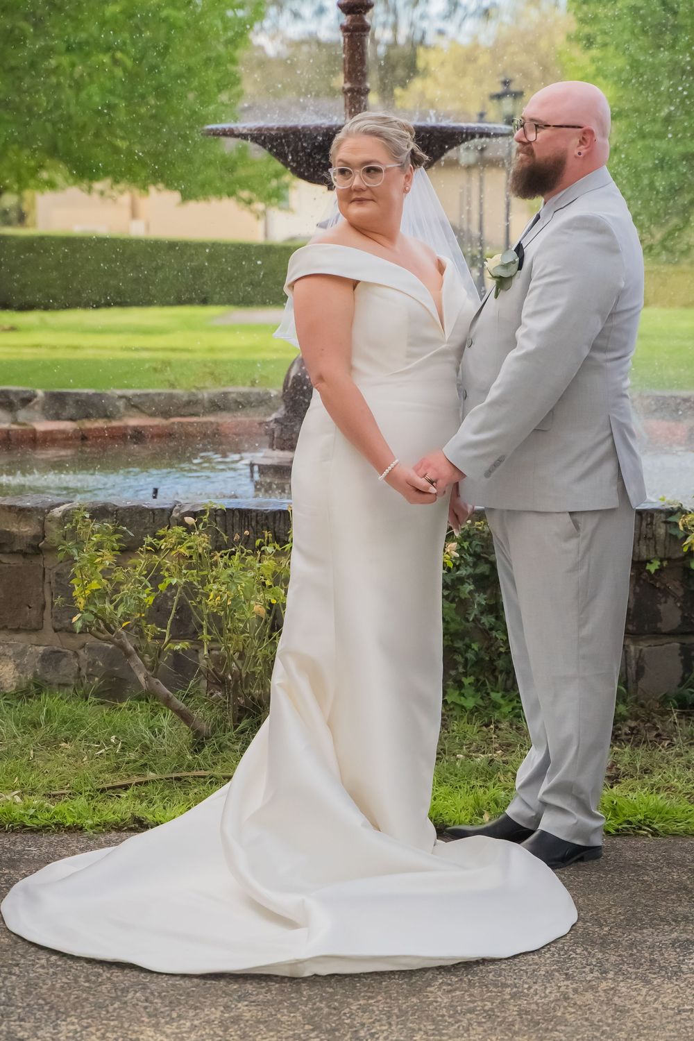 A couple in formal wedding attire poses together by a tranquil pond, showing details of the bride's flowing white dress train.