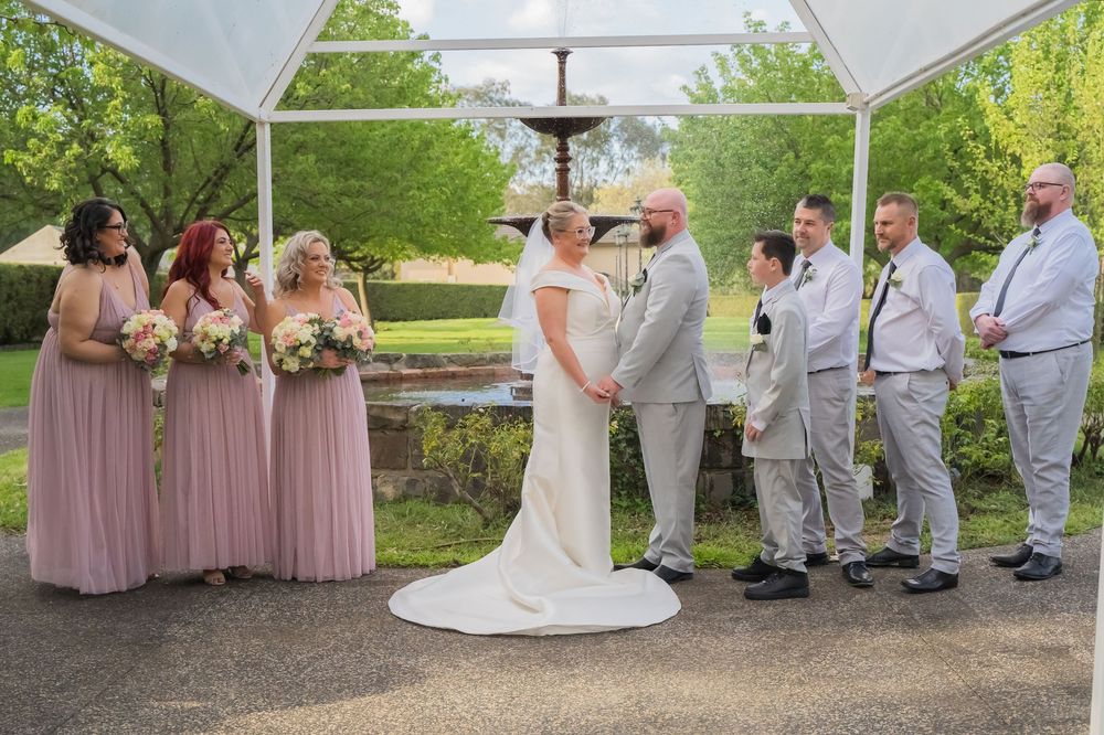 Wedding party poses for photos under outdoor pavilion with bridesmaids in pink dresses and groomsmen in light gray suits.