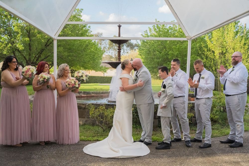 Wedding ceremony takes place under a white canopy outdoors with bridesmaids in pink dresses and groomsmen in gray suits.