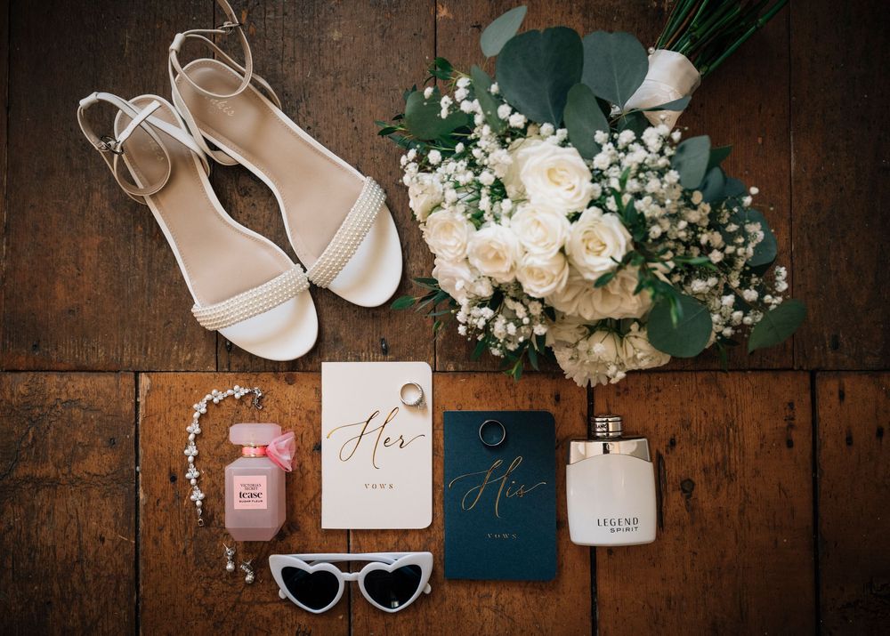 Wedding accessories including white heels, perfume, invitations and floral bouquet arranged on wooden surface.