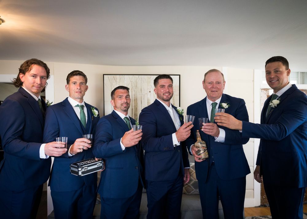 Groomsmen in navy suits toast with drinks during pre-wedding preparations.