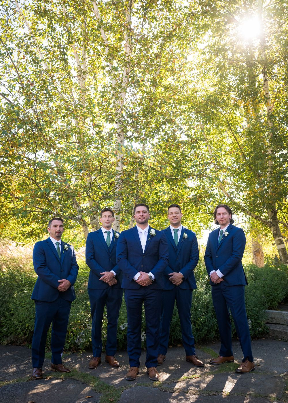 Groomsmen in navy suits pose for photos in a sunlit outdoor setting.