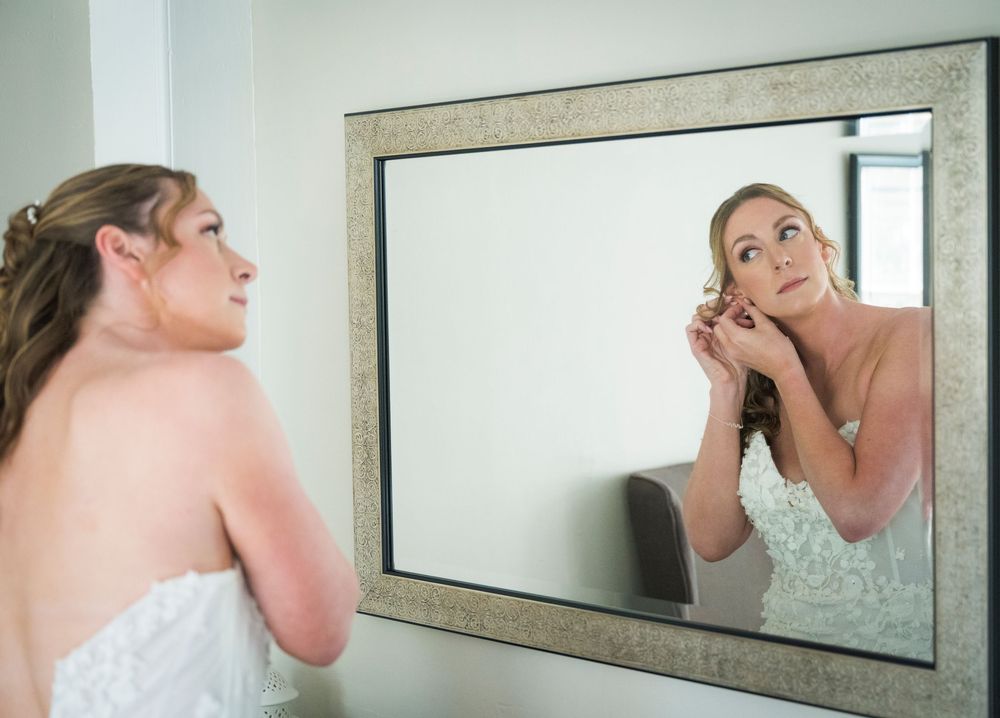 Bride getting ready in front of mirror wearing white lace wedding dress.