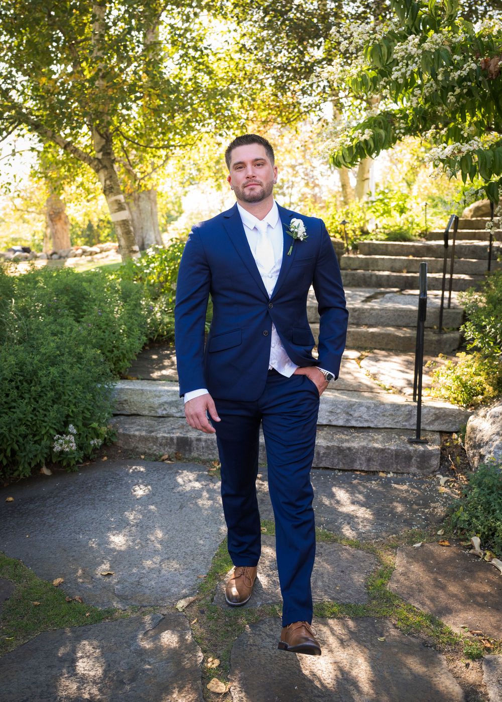 Navy blue suited figure poses in dappled sunlight on garden path.