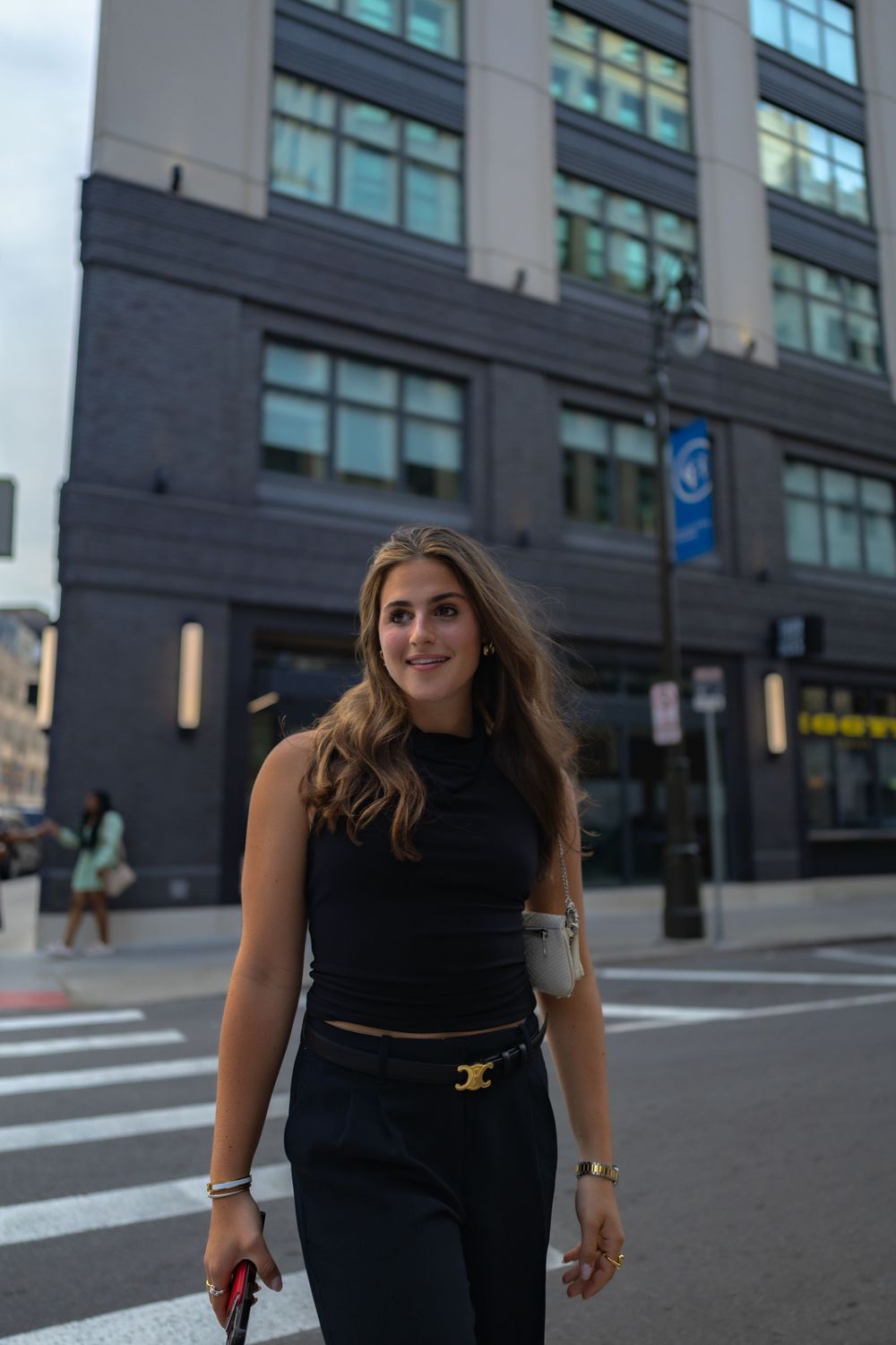 A person in all black clothing walks down a city sidewalk with modern office buildings in the background.