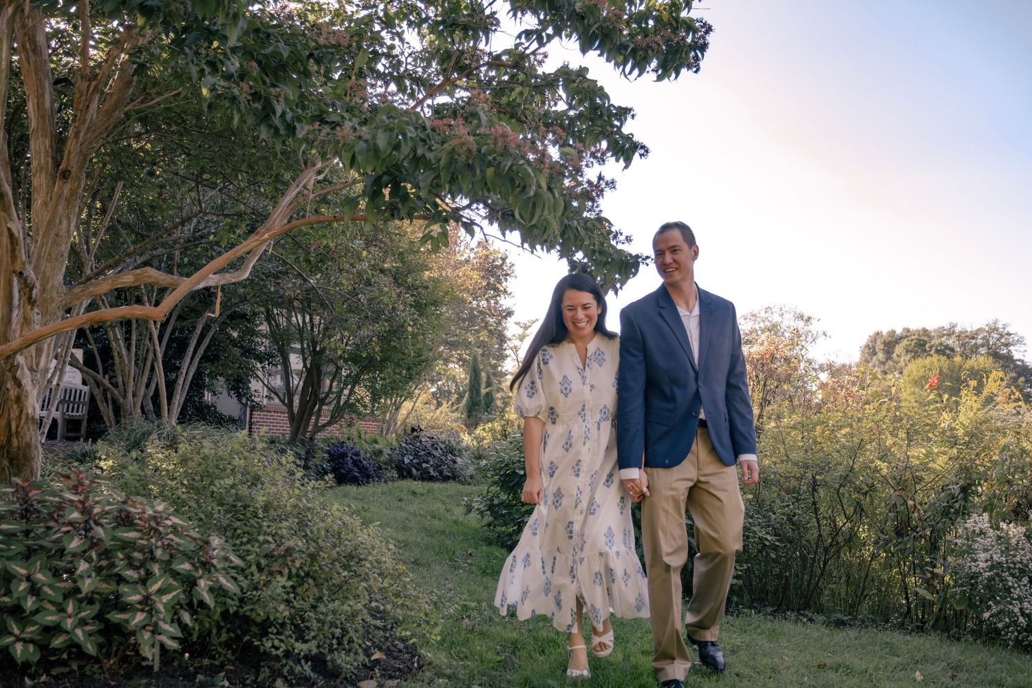 Couple in formal attire stands together in a garden setting during golden hour lighting.