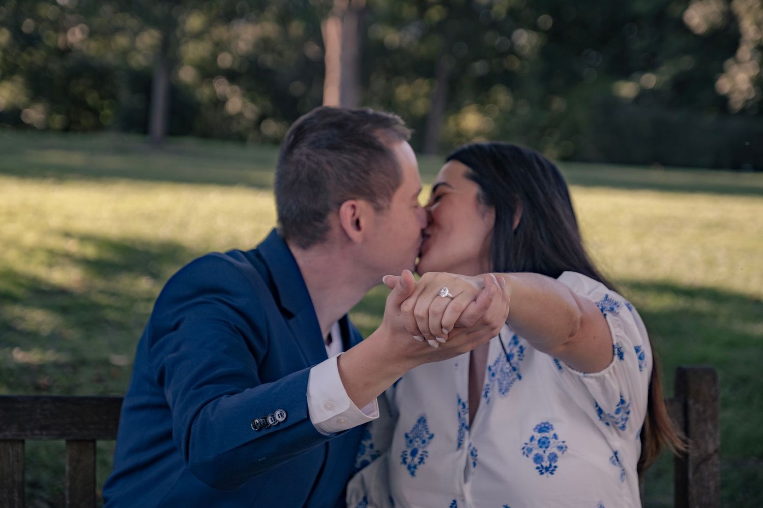A romantic couple shares a kiss while sitting on a wooden bench in a sunny outdoor park setting.