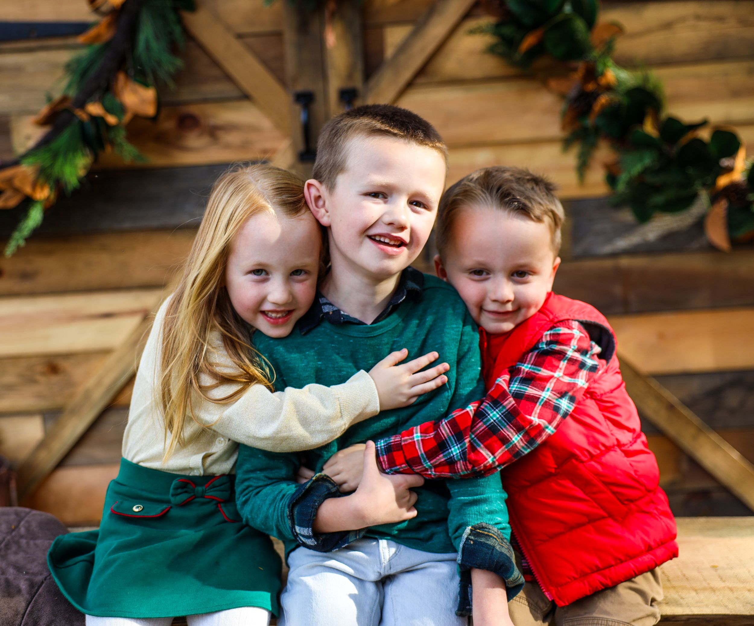 Three children sharing warm embrace while sitting on wooden bench with holiday decorations.
