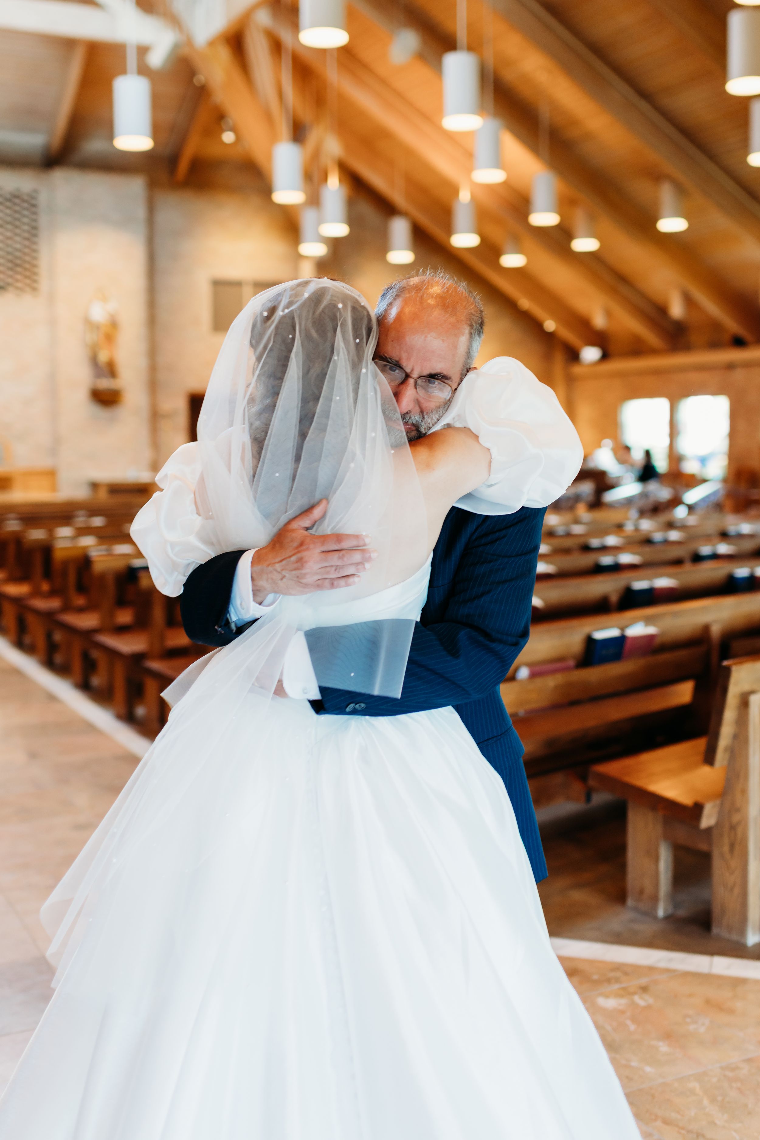 A touching moment shared between two people on their wedding day inside a church with wooden pews and pendant lights.