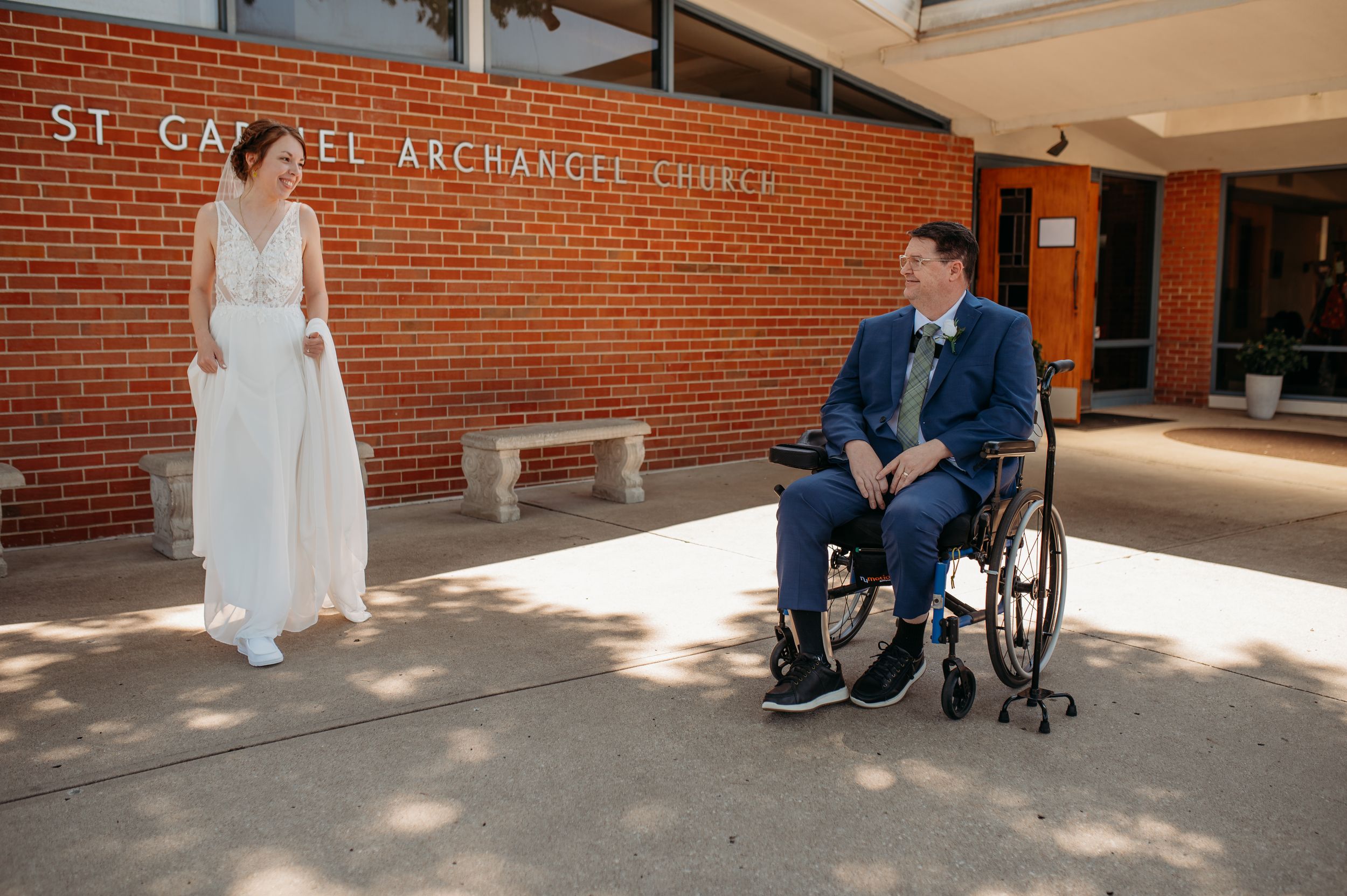 Two people stand in front of St. Gabriel Archangel Church, with one person seated in a wheelchair.