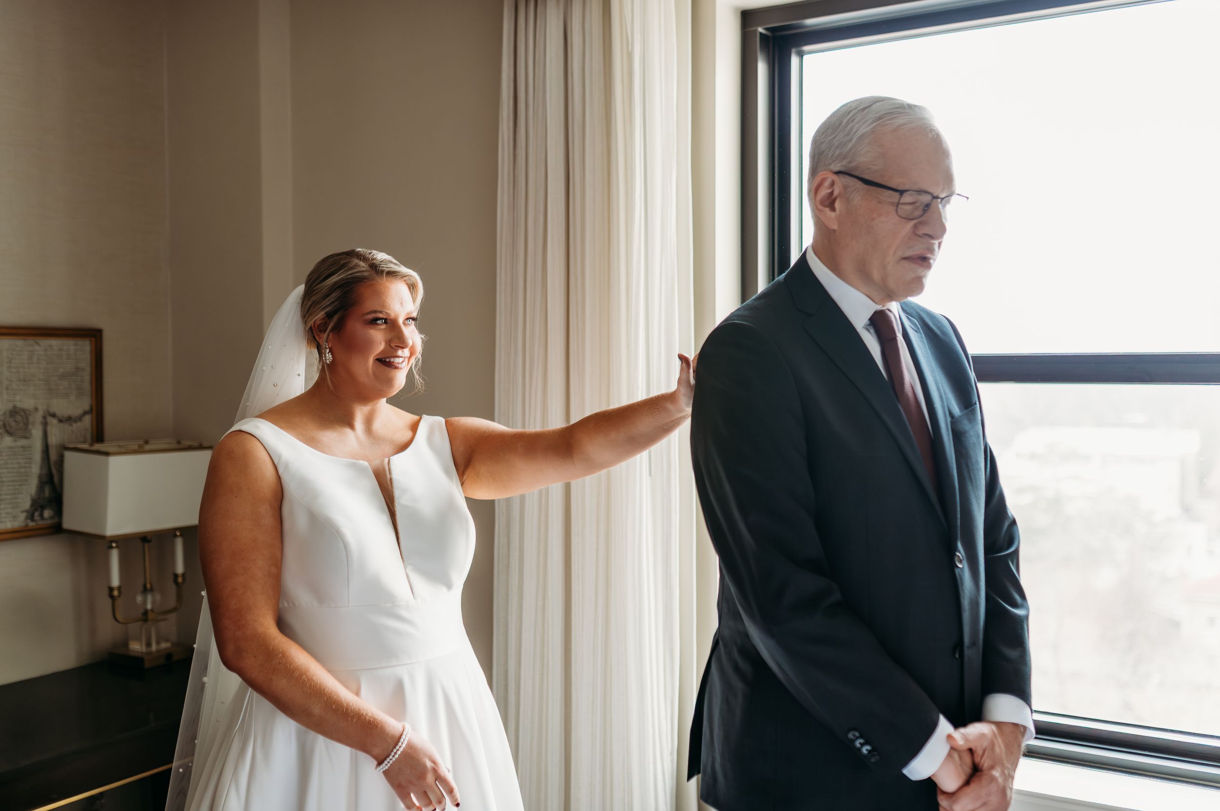 A touching moment between a bride in a white dress and someone in a dark suit by a window.