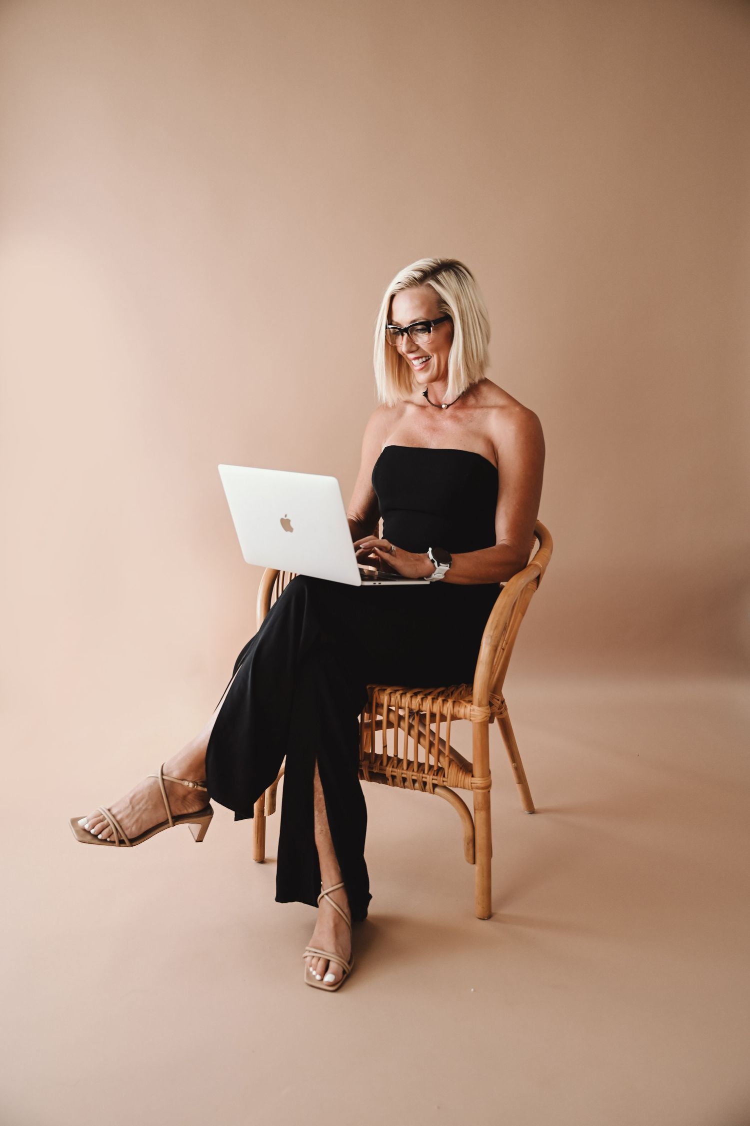 Woman in black dress poses on rattan chair against peachy pink studio backdrop.