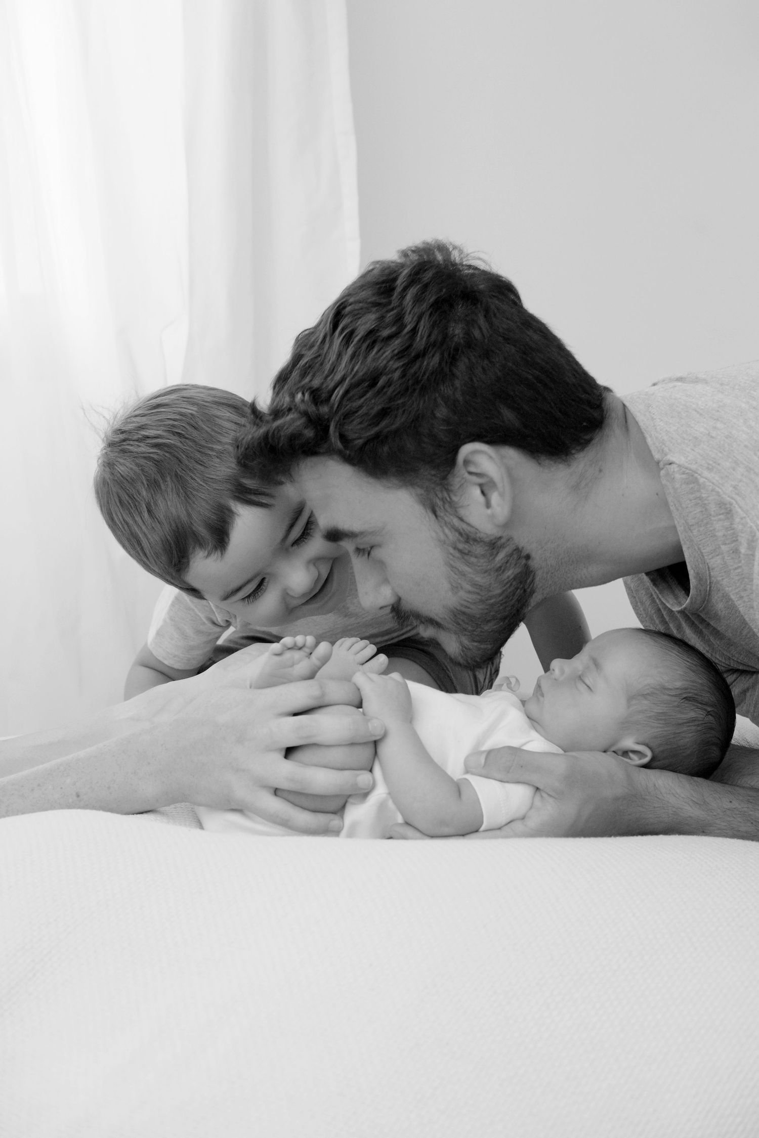 Fotografía en blanco y negro que muestra la emoción de un padre y su hijo de tres años besando a su bebé recién nacido