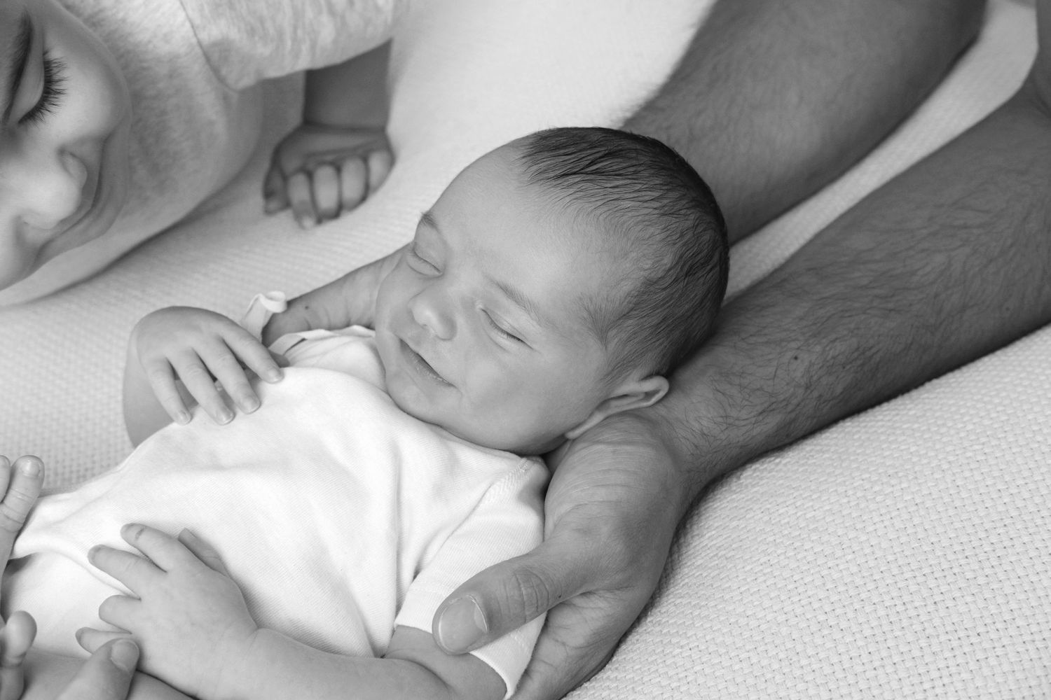 Un bebé recién nacido sonríe placenteramente mientras duerme en brazos de su padre y su hermano le observa de reojo