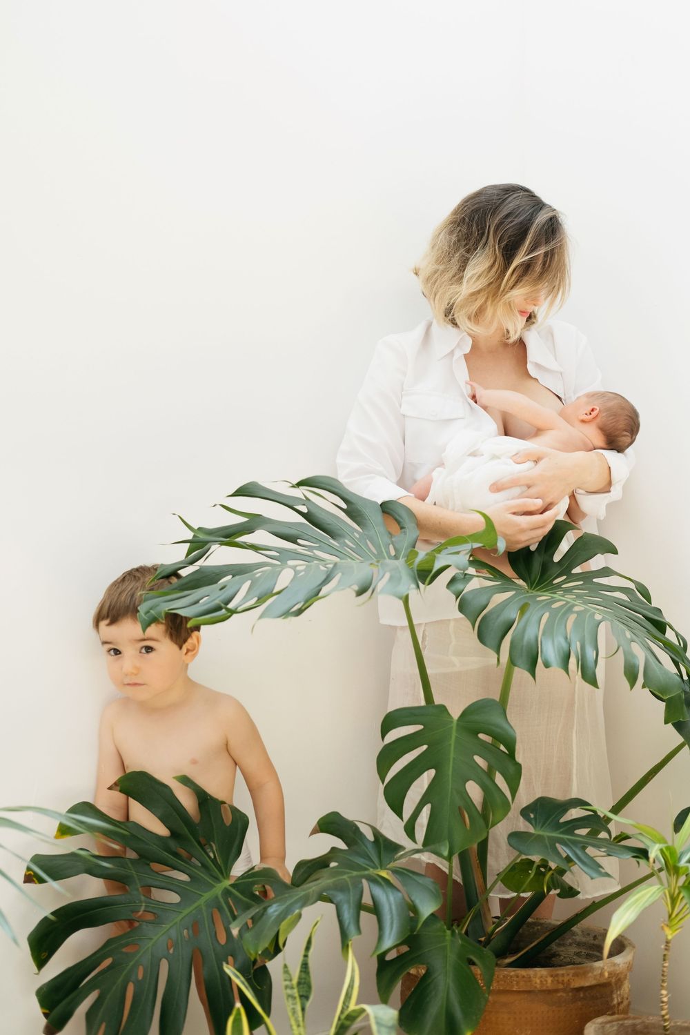 Imagen familiar de una madre dando de mamar a su bebé recién nacido junto a su hijo mayor que se esconde tras una planta monstera deliciosa